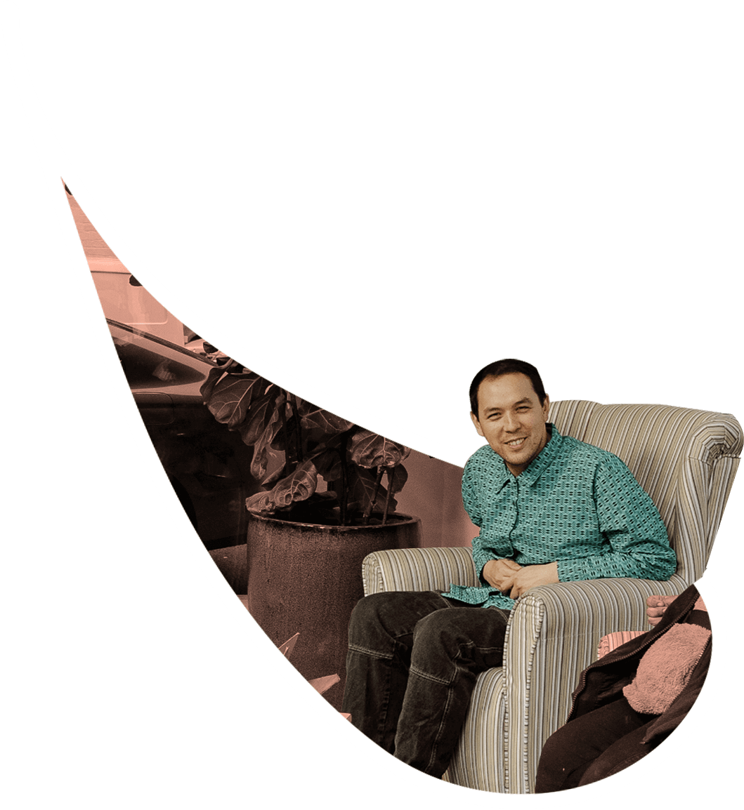 A young person wearing a green patterned shirt and jeans sits in an armchair, leaning forward keenly and smiling at the camera.