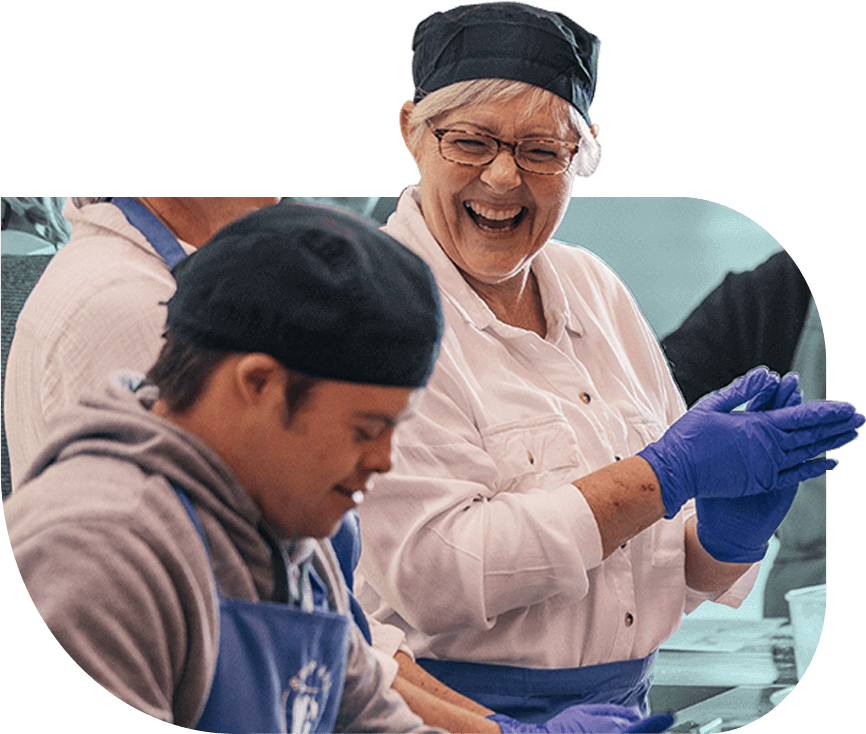 Two bakers from social enterprise Step and Stone smile together in a commercial kitchen. They wear blue gloves and aprons and black chef caps.