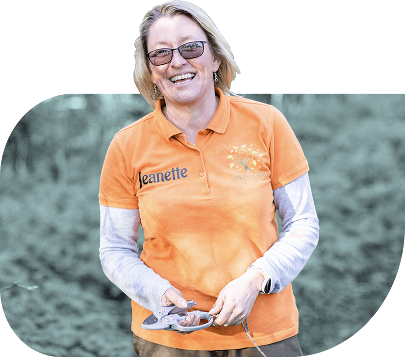 A blonde social entrepreneur smiles outdoors wearing an orange t-shirt and holding a branch and a plant trimming tool.