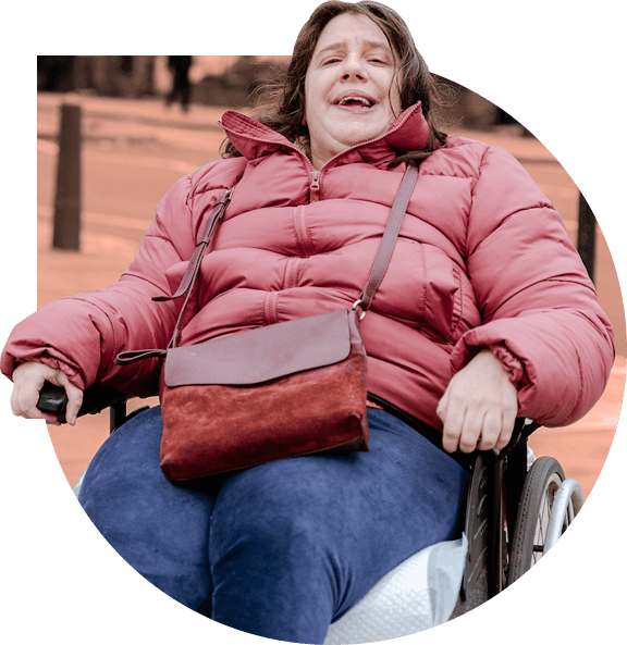 Anna Alston, a member of Camden Disability Action social enterprise smiles, using a wheelchair and wearing a red mauve puffer coat with a matching handbag.